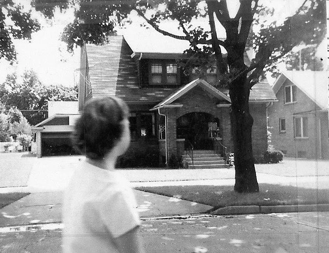 Black-and-white photo of a blurred child in the foreground looking toward a two-story brick house with a front porch and a large street-tree.
