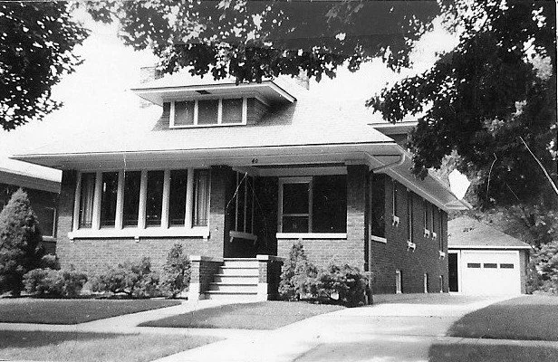 Black-and-white photo of a brick bungalow with front steps, a covered porch, detached garage, and trees framing the yard.