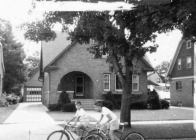 Black-and-white photo of two boys with bicycles on a sidewalk in front of a brick bungalow with an arched porch and a large tree.