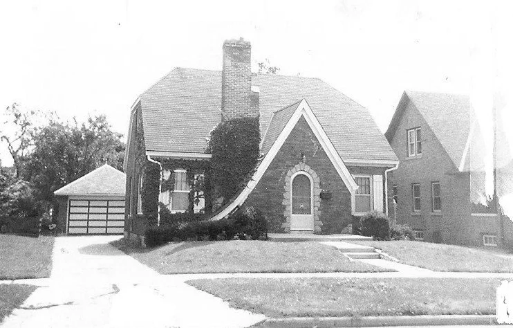 Black-and-white photo of a brick Tudor-style house with a steep gabled roof, arched front door, ivy-covered chimney, front lawn and driveway leading to a detached garage.