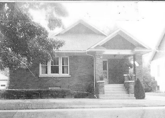 Black-and-white photo of a single-story brick bungalow with a front porch, steps, and a large tree on the left.
