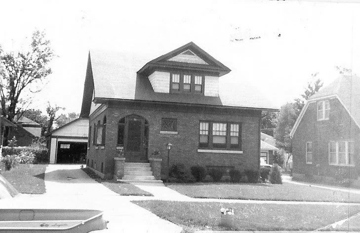 Black-and-white photo of a brick two-story house with a front porch and steps, dormer window, driveway leading to a detached garage, and a lawn with a parked car partially visible at left.