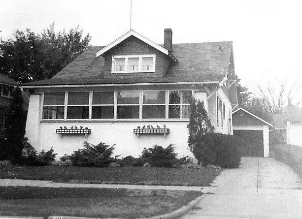 Black-and-white photo of a single-family bungalow with an enclosed front porch, shrubs in the yard, and a driveway leading to a detached garage.