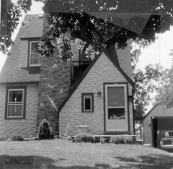 Black-and-white photo of a single-family bungalow with an enclosed front porch, shrubs in the yard, and a driveway leading to a detached garage.