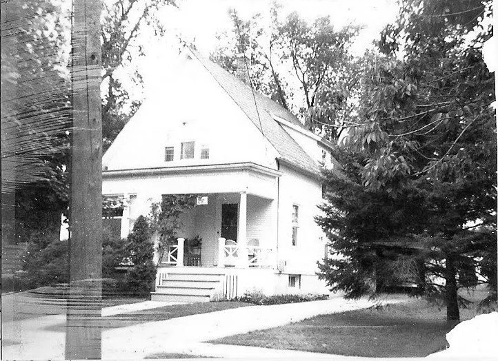 Black-and-white photo of a two-story house with a covered front porch and steps, surrounded by trees and a sidewalk in the foreground.