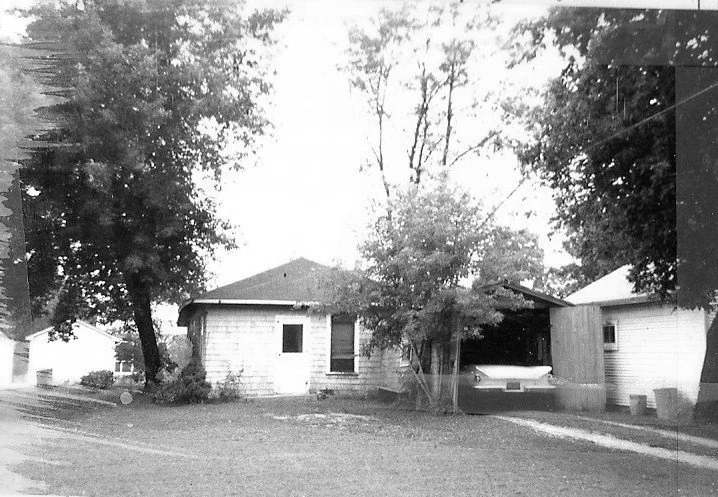 Black-and-white photo of a single-story house with a carport sheltering a vintage car, trees in front and a grassy yard.