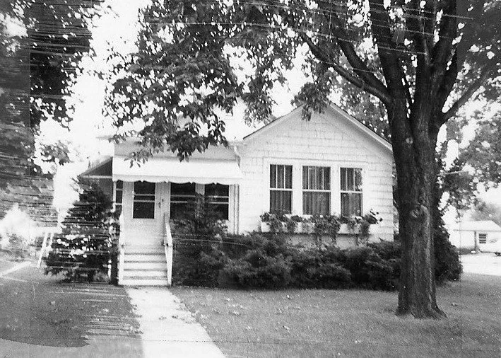Black-and-white photo of a small single-story house with a front porch, shrubs, a large tree in the yard, and a sidewalk leading to the steps.