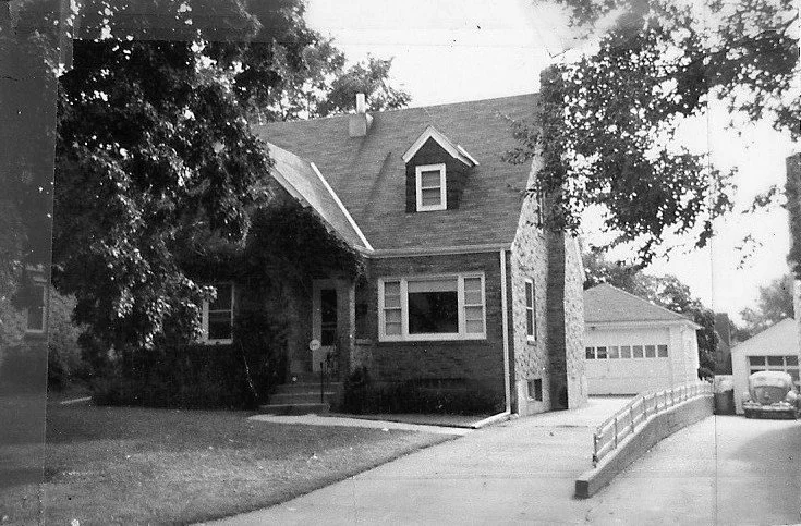 Black-and-white photo of a brick two-story house with a dormer and front porch, driveway leading to a detached garage and a vintage car.
