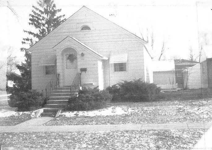 Small single-story house with front steps and arched porch, shrubs in front yard, light snow on the lawn and a detached garage to the right.
