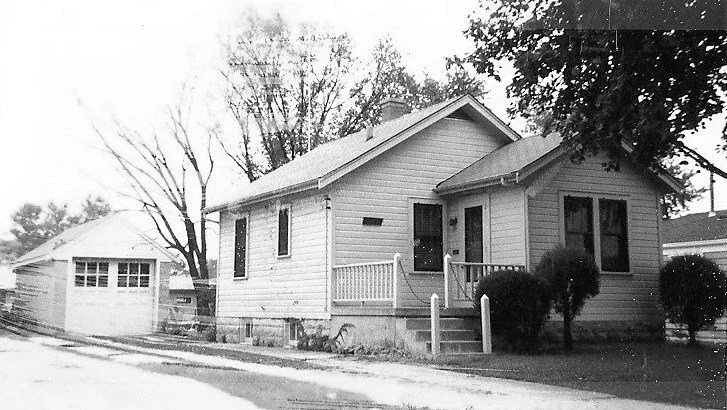 Black-and-white photo of a small single-story house with front steps and porch, detached garage, and trees.