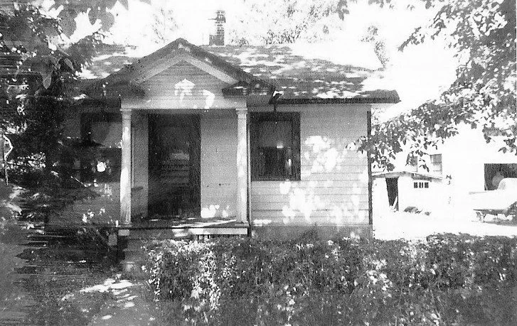 Black-and-white photo of a small single-story house with a front porch, columns, shrubs, and dappled tree shadows; garage and car visible to the right.
