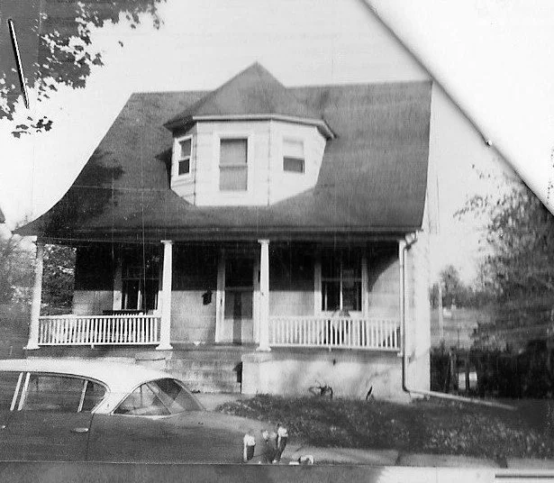 Black-and-white photo of a two-story house with a front porch, octagonal dormer window, and a parked car in the foreground.