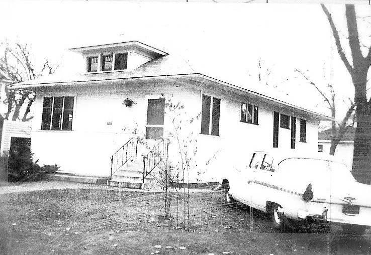 Black-and-white photo of a single-story bungalow with front steps and railing, a vintage car parked on the right, and leafless trees in the yard.