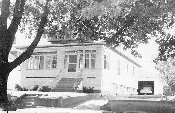 Black-and-white photo of a single-story bungalow with an enclosed front porch, steps up to the door, a large tree in the foreground, and a driveway leading to a garage with a car.
