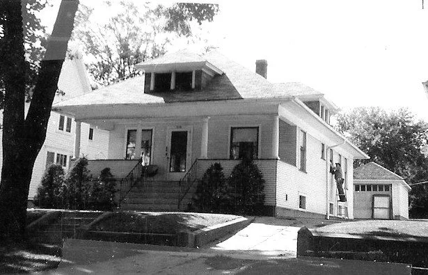 Black-and-white photo of a single-story bungalow with a front porch, stairs and shrubs, a detached garage at right, and a large tree in the foreground.
