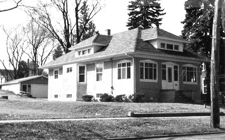 Black-and-white photo of a single-story bungalow-style house with an enclosed front porch, detached garage to the left, and leafless trees in the yard.
