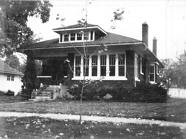 Black-and-white photo of a single-story Craftsman-style bungalow with front steps, a small tree in the yard, and a tall chimney.