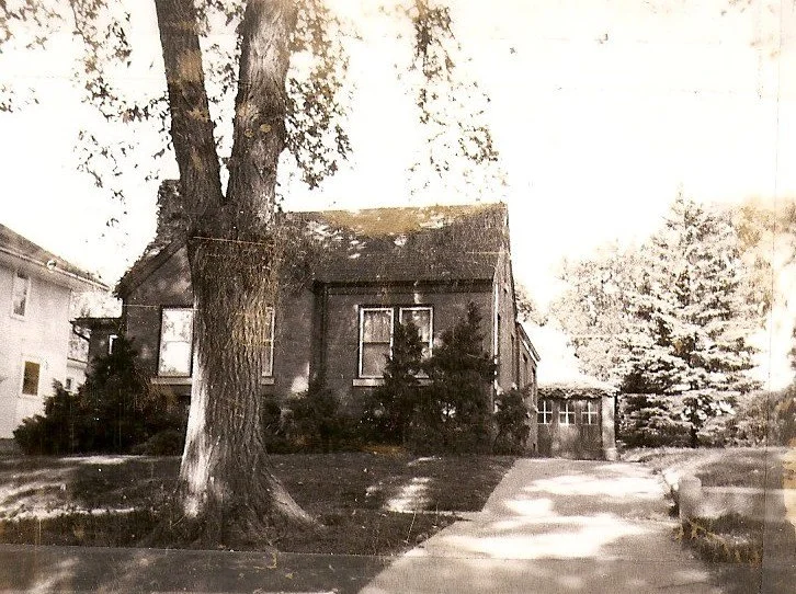 Sepia photo of a small brick house with a large tree in the front yard, a sidewalk and driveway leading to a detached garage.