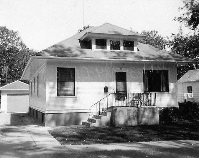 Black-and-white photo of a single-story bungalow with a front porch and steps, dormer window on the roof, detached garage to the left, and trees in the background.