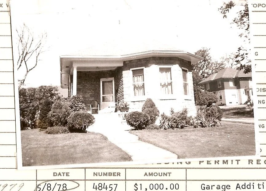 Black-and-white photo of a single-story brick bungalow with a bay window, front steps, shrubs and lawn.