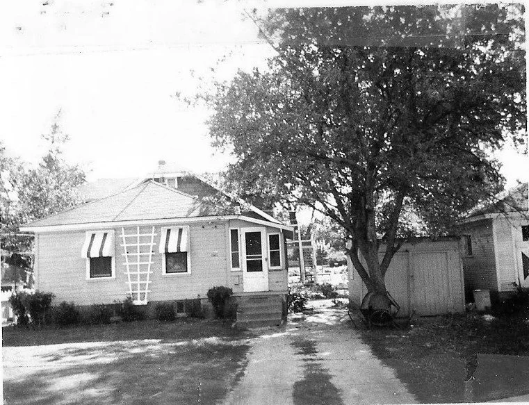 Black-and-white photo of a small one-story house with striped window awnings, front steps, driveway, large tree and a shed to the right.