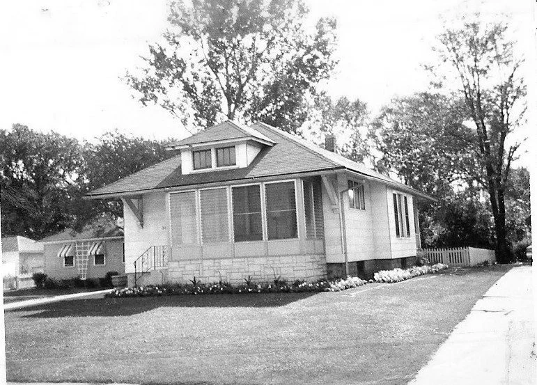 Black-and-white photo of a single-story bungalow with an enclosed front porch, lawn, and trees.