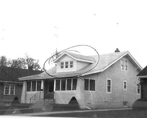 Black-and-white photo of a single-story bungalow with enclosed front porch and a circled dormer window on the roof.