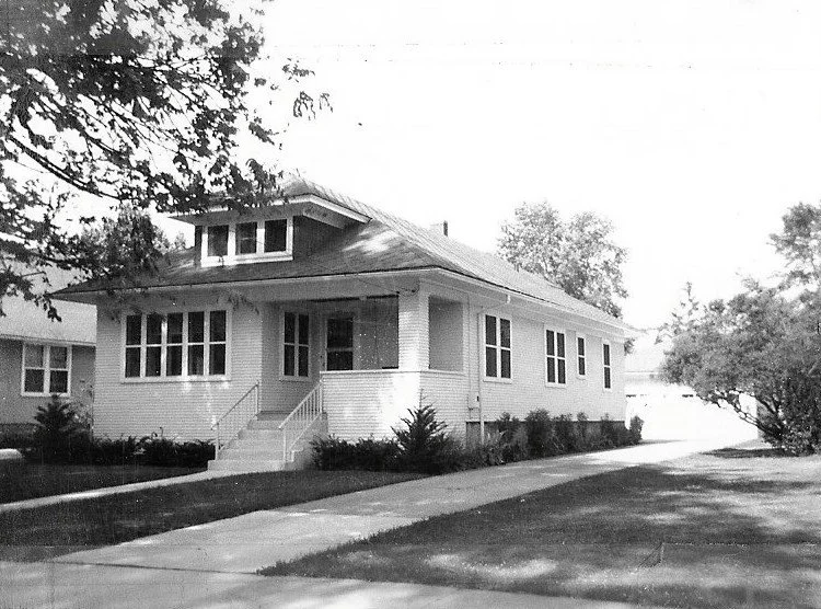 Black-and-white photo of a single-story bungalow with a covered front porch, steps leading up, multiple windows, driveway to the right, and trees/lawn in the yard.