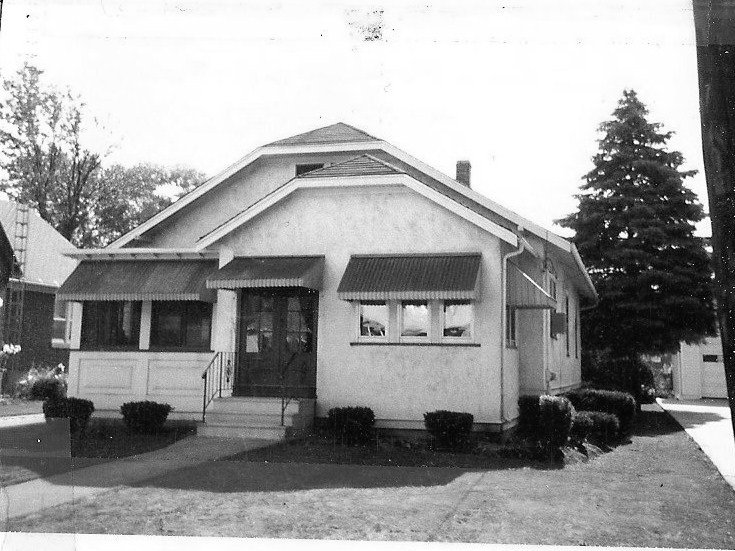 Black-and-white photo of a small single-story bungalow with a covered front porch, window awnings, low shrubs, and a driveway with a tall evergreen at the right.