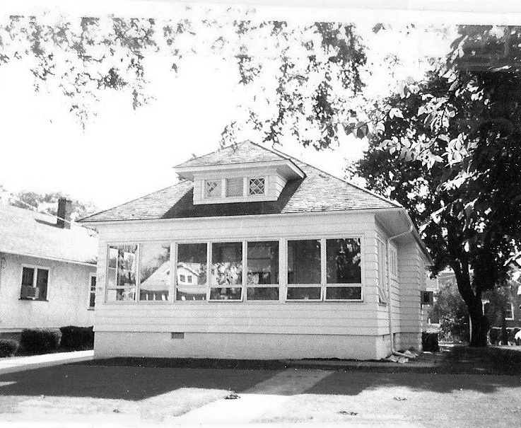 Black-and-white photo of a one-story bungalow with an enclosed glass front porch, small dormer window, and trees framing the house.