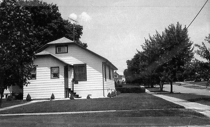 Black-and-white photo of a small single-story house on a corner lot with a front stoop, manicured lawn, sidewalk, and trees lining the street.