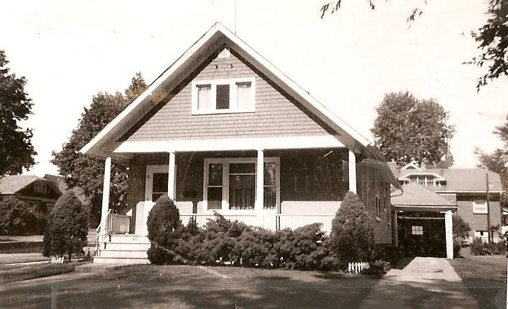 Sepia-toned front view of a single-story bungalow with a covered porch, shrubs, and a side driveway.