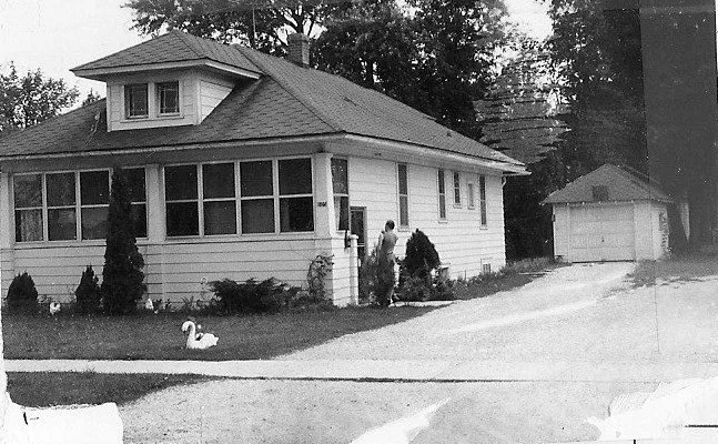 Black-and-white photo of a one-story bungalow with an enclosed front porch, a person at the side door, a swan lawn ornament on the front yard, and a detached garage at the end of the driveway.