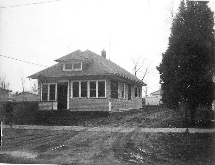 Black-and-white photo of a single-story bungalow with an enclosed front porch, dirt driveway, and a large tree at the right.