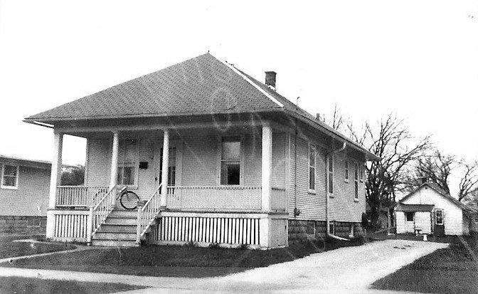 Black-and-white photo of a single-story bungalow with a covered front porch, steps, driveway, and a detached garage.