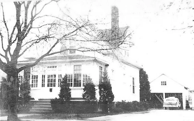Black-and-white photo of a white two-story house with an enclosed porch, bare front tree, and a driveway leading to a garage with a parked car.