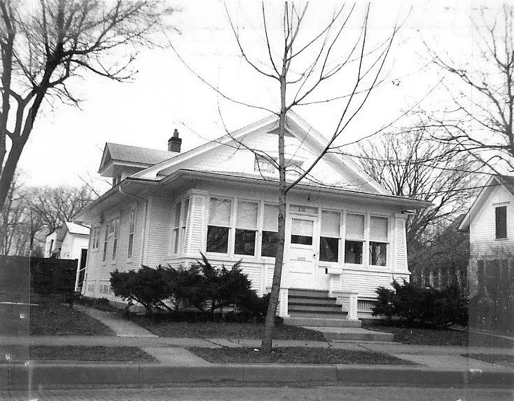 Black-and-white photo of a one-story bungalow with an enclosed front porch, front steps, leafless tree and shrubs, and a sidewalk.