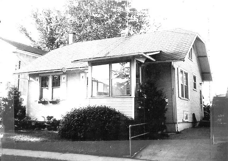 Black-and-white photo of a one-story bungalow with a front bay window, low shrubs, and a sidewalk in front.