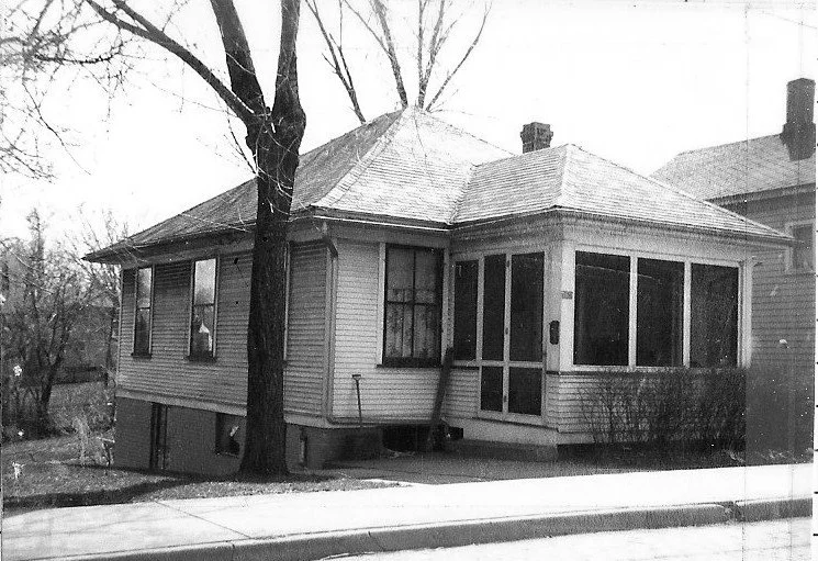 Black-and-white photo of a single-story wood-frame house with a screened front porch, a large tree at the front corner, and a sidewalk in the foreground.