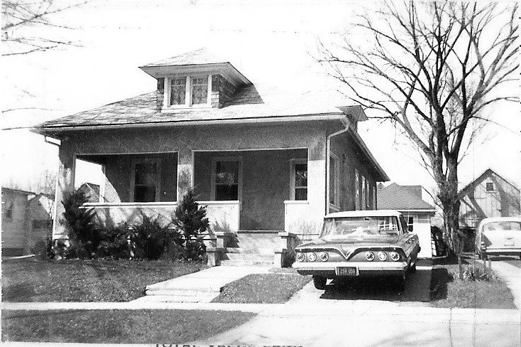 Black-and-white photo of a single-story bungalow with a covered front porch, a vintage car parked in the driveway, and a leafless tree at the right.
