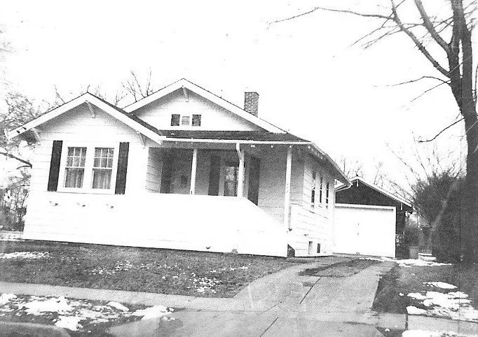 Black-and-white photo of a single-story bungalow with a covered front porch, driveway leading to a detached garage, leafless trees, and small patches of snow on the lawn.