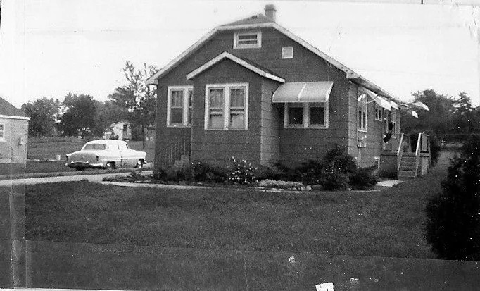 Black-and-white photo of a small single-story bungalow with awnings, front lawn and shrubs, and a vintage car parked on the street.