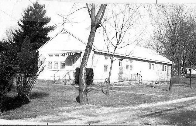 Black-and-white photo of a single-story bungalow with a front porch and awning, surrounded by leafless trees and a grassy yard.