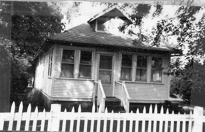 Black-and-white photo of a small single-story bungalow with a screened front porch, short steps up to the door, and a white picket fence in the foreground.