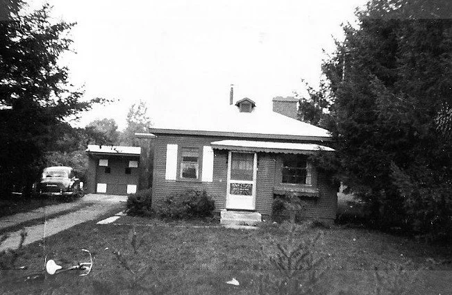 Black-and-white photo of a small brick bungalow with a front porch and awning, a driveway leading to a detached garage with a vintage car parked nearby, and trees framing the yard.