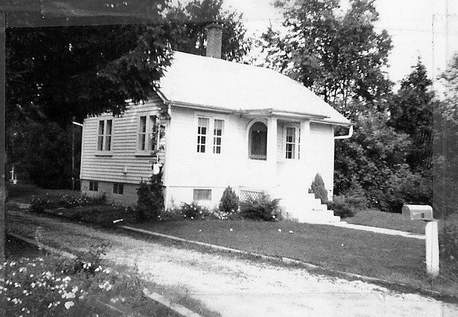 Black-and-white photo of a small single-story bungalow with front steps, lawn, mailbox, and trees in the background.