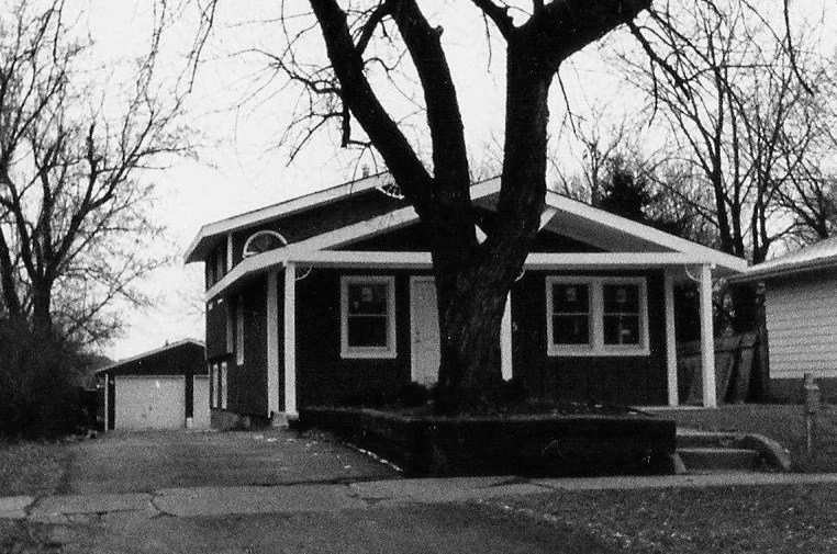 Black-and-white photo of a single-story bungalow with a large tree in front and a driveway leading to a detached garage.