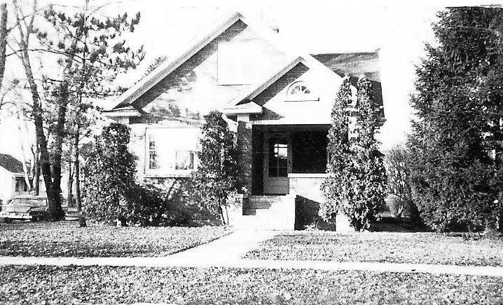Black-and-white photo of a bungalow-style house with a front porch, steps, tall shrubs on either side, and a sidewalk and lawn in front.