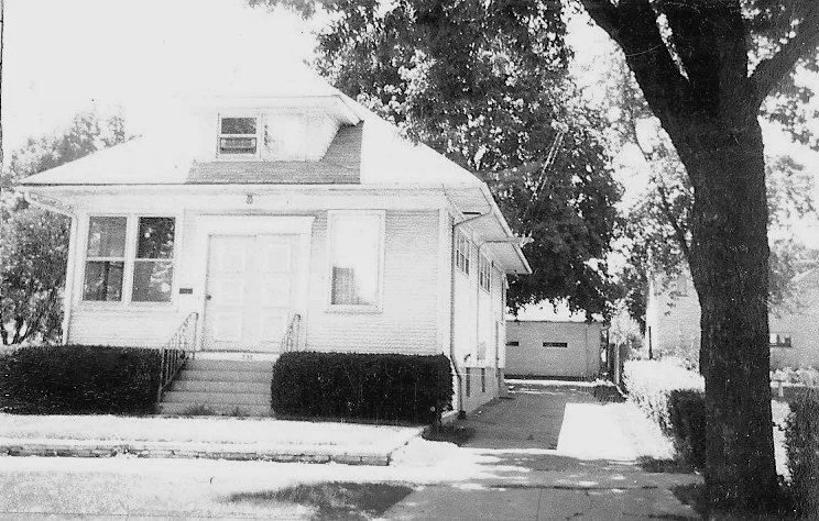 Black-and-white photo of a small two-story bungalow with front steps and shrubs, a driveway to a detached garage, and a large tree in the foreground.