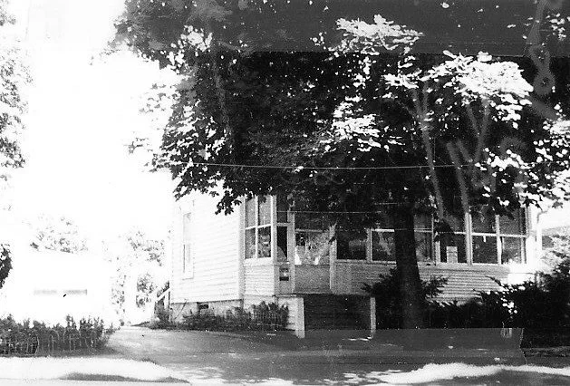 Black-and-white photo of a two-story house with an enclosed front porch, a large tree shading the facade, and a driveway in front.